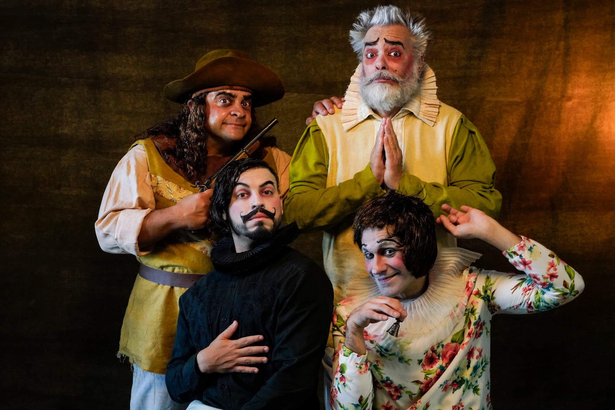 Quatro artistas caracterizados com figurinos de época, em retrato de estúdio, com expressões cômicas e frontalidade de farsa.