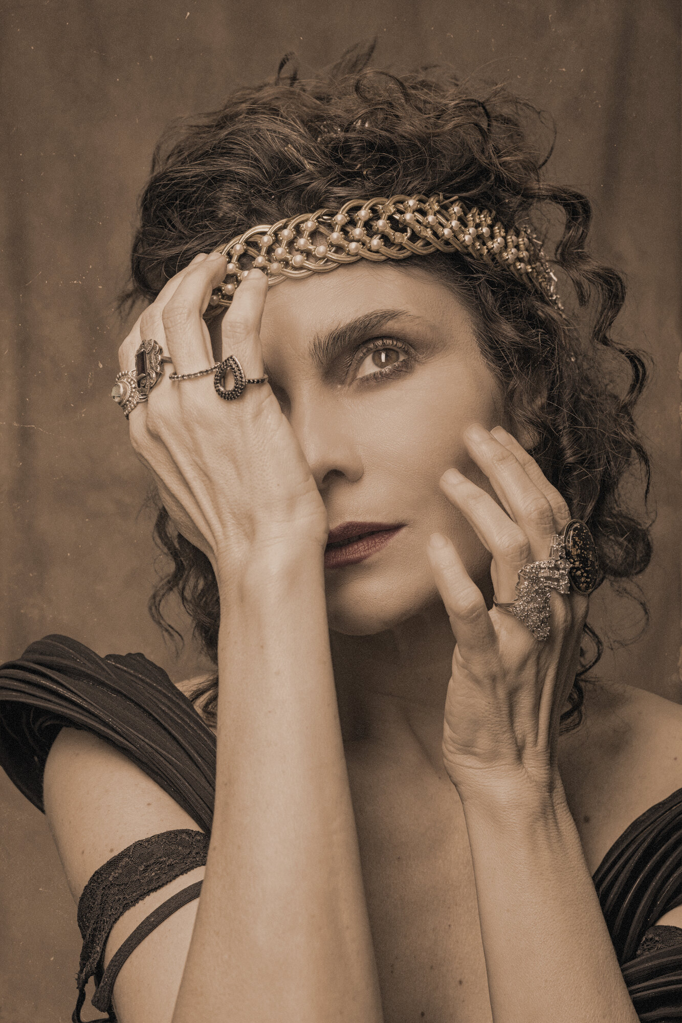 A atriz que faz Sarah Bernhardt com uma das mãos cobrindo parte do rosto, tiara de pérolas e anéis