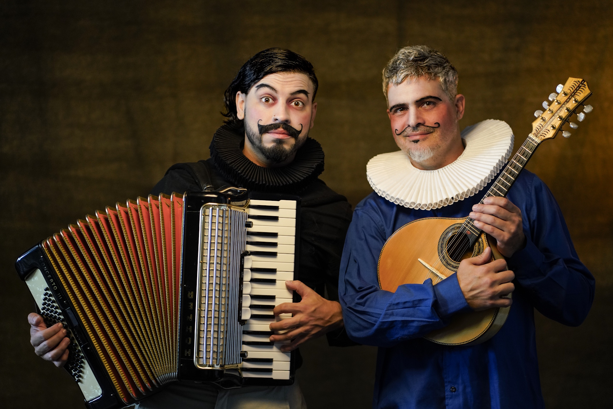 Dois artistas caracterizados; um segura um acordeon e o outro um instrumento de corda, em retrato de estúdio.