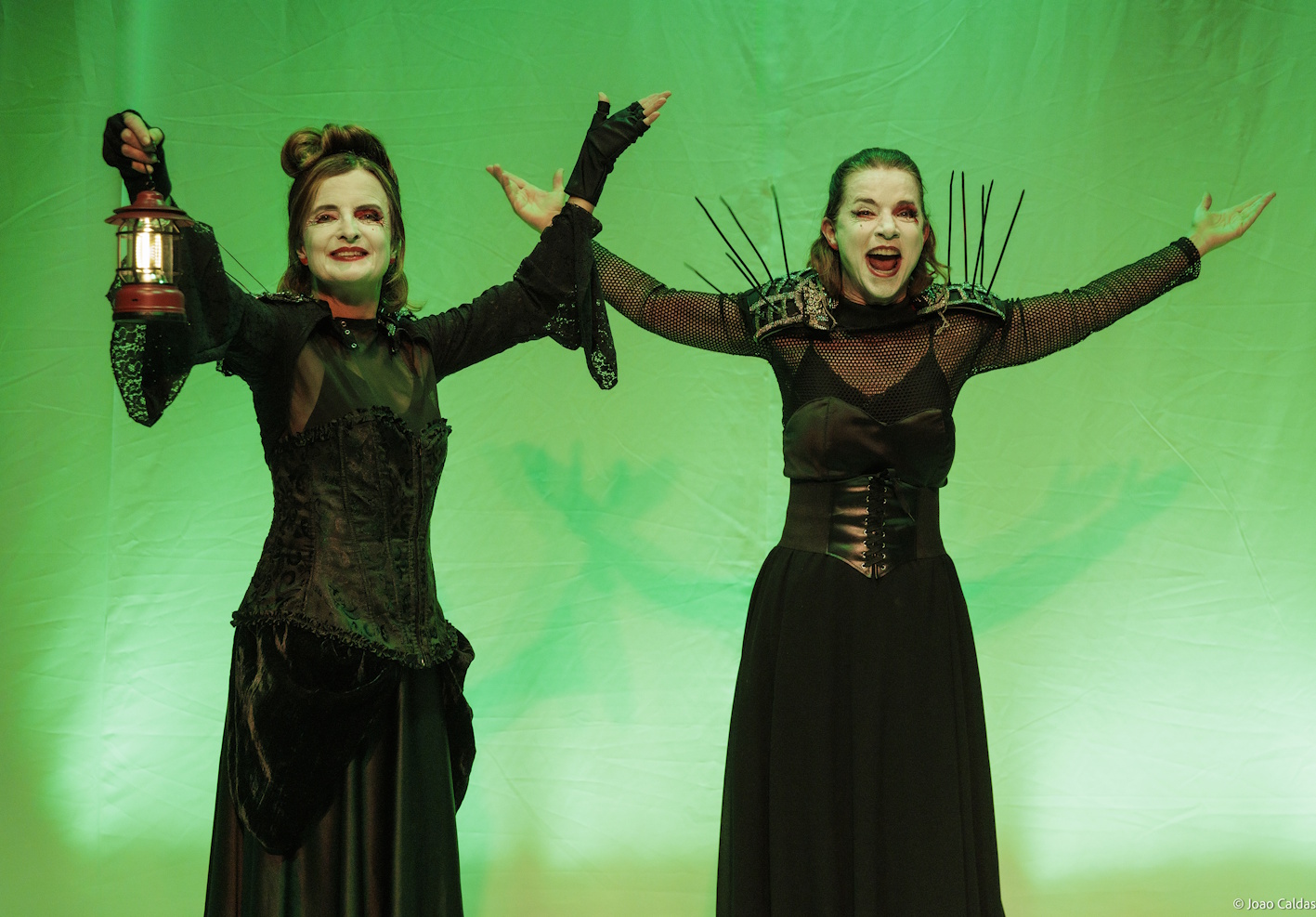 Duas intérpretes em luz verde; uma segura uma lanterna; clima de jogo teatral e humor.