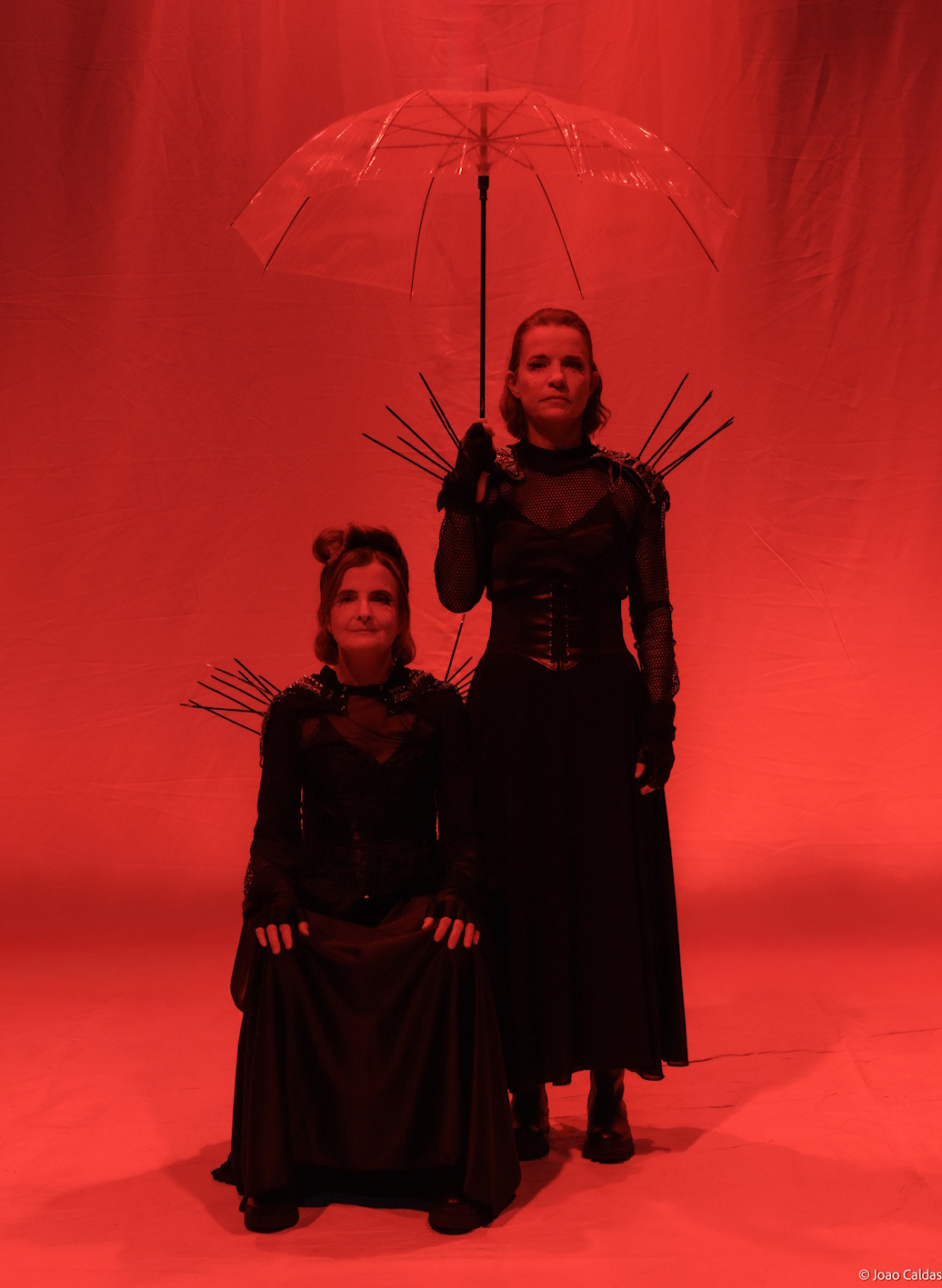 Duas intérpretes em luz vermelha; uma sentada, outra em pé segurando guarda-chuva transparente; figurinos pretos.