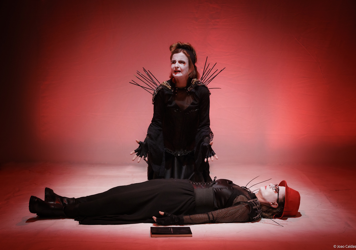 Tableau em luz vermelha: uma intérprete ajoelhada em gesto ritual, outra deitada; atmosfera trágica.