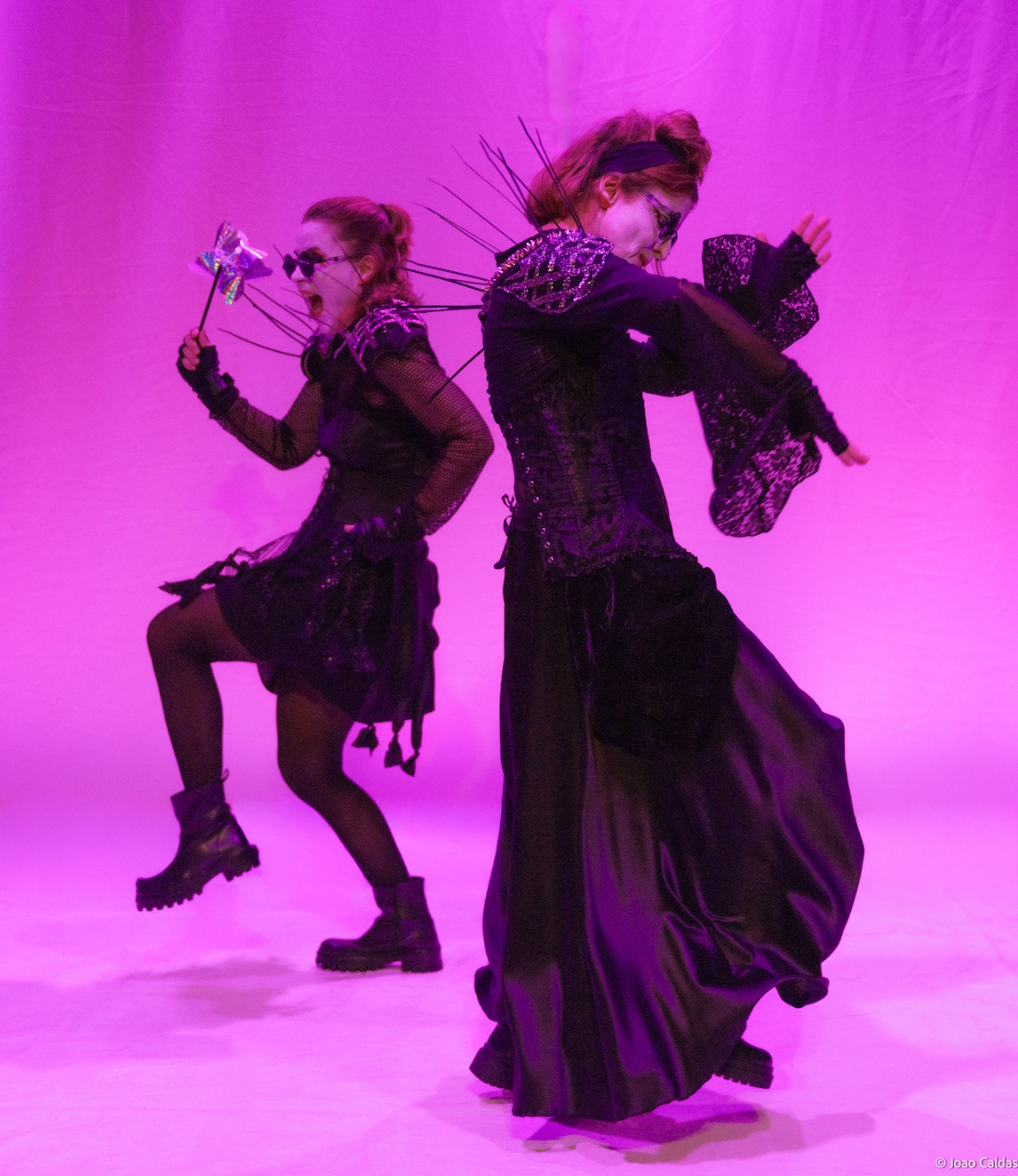 Duas intérpretes em luz magenta, em movimento; uma gira com vestido longo, a outra em gesto cômico.