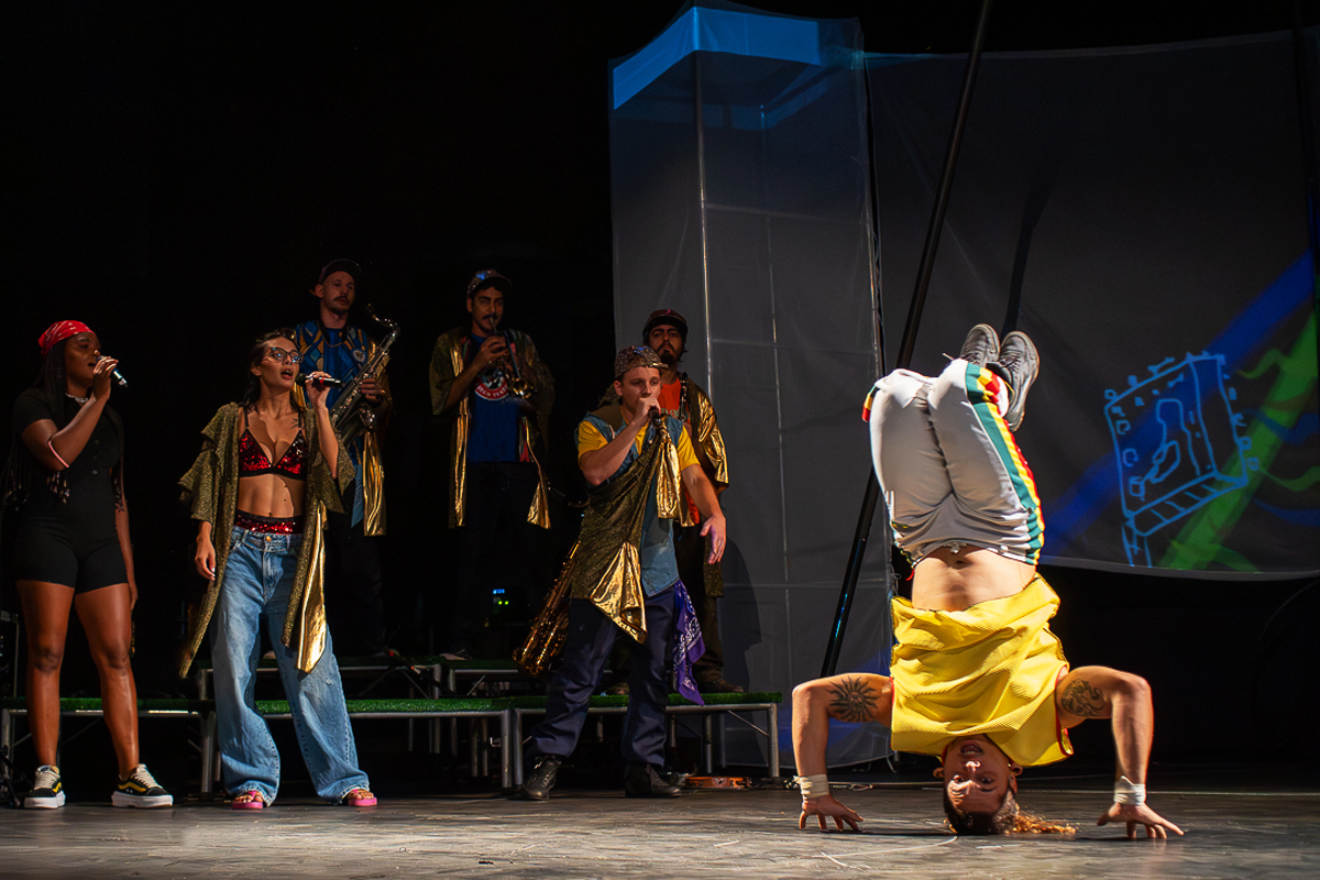 Mistura de música ao vivo e acrobacia: performer em posição invertida com cantoras e músicos ao fundo.