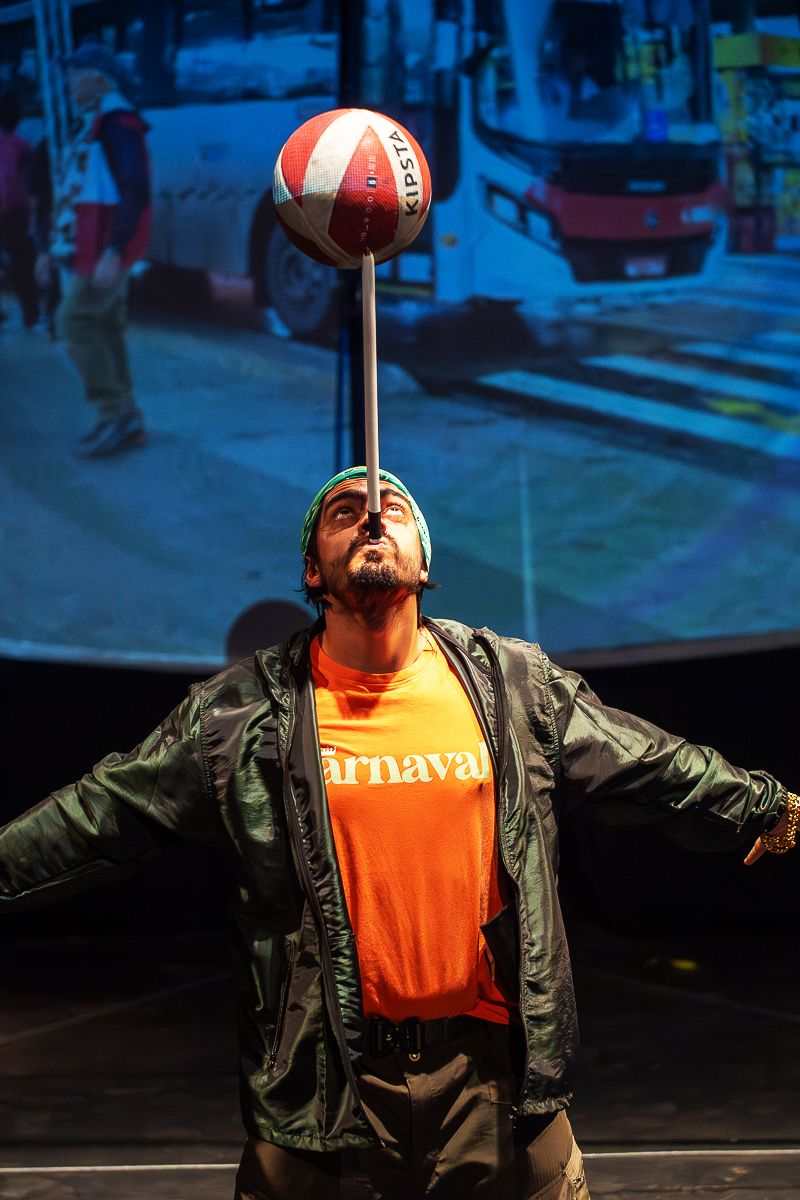 Número circense em close: performer equilibrando bola em haste, com projeção ao fundo.