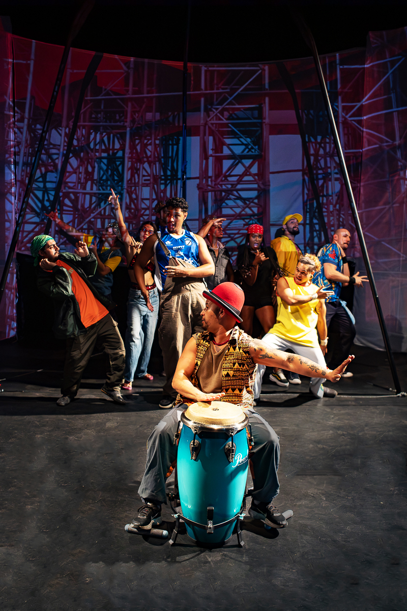 Coro e ritmo em cena: tambor em primeiro plano e performers ao fundo em composição coletiva.