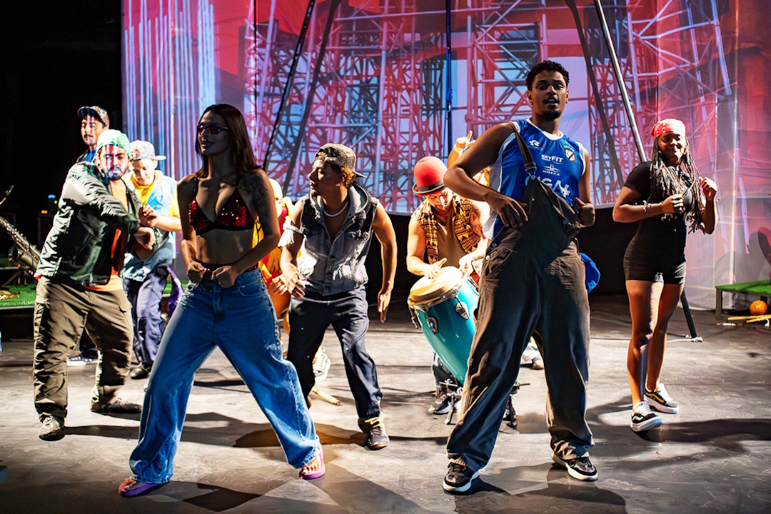 Cena de conjunto: performers em movimento, música e dança em cena.