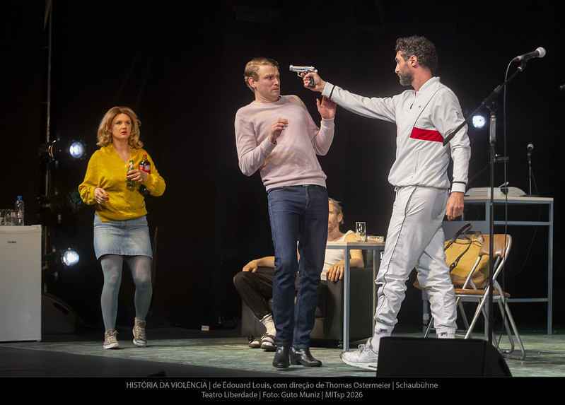 Ator aponta arma para a cabeça de outro, atriz ao fundo com refrigerante