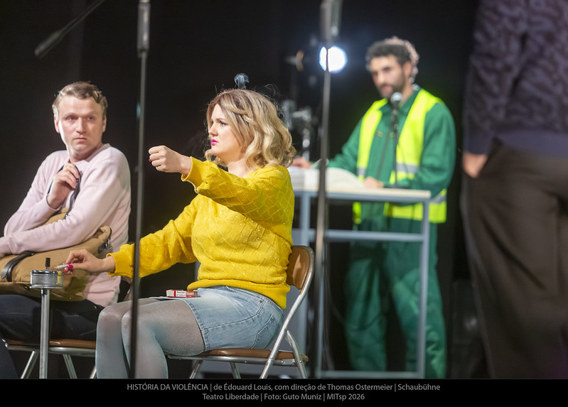 Atriz de suéter amarelo com cigarro, ator ao lado, homem de colete verde ao fundo