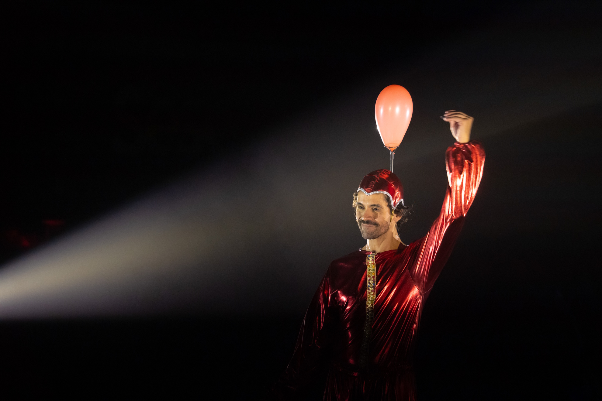 Ator caracterizado como um pin de mapa com figurino vermelho e balão vermelho sobre a cabeça em Pés-Coração.
